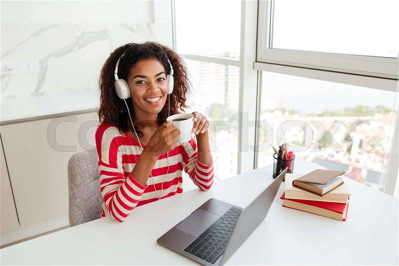 Smiling african woman sitting by the table with laptop computer, drinking coffee and looking at the camera, stock photo