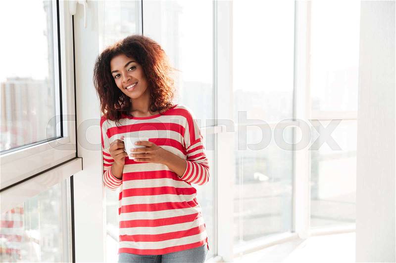 Happy african woman drinking coffee near the window and looking at the camera, stock photo