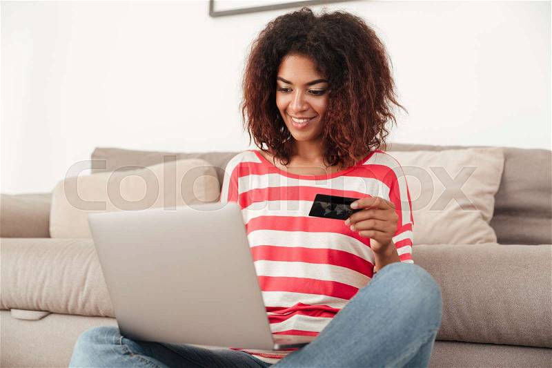 Smiling african woman sitting on bed with credit card and laptop computer at home, stock photo