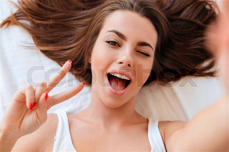 Close up portrait of a pretty young woman winking while making a selfie in bed, stock photo