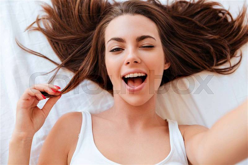 Close up portrait of a cheerful young woman winking while making a selfie in bed, stock photo
