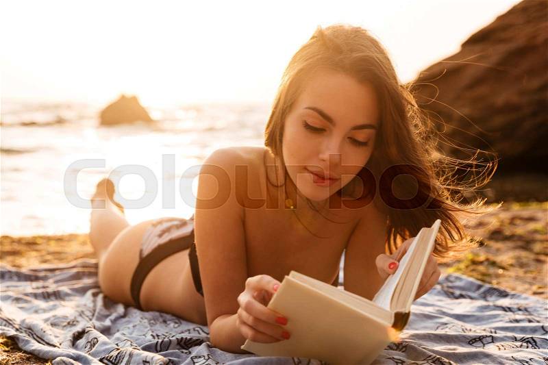 Pretty brunette woman in bikini lying on beach and reading book, stock photo