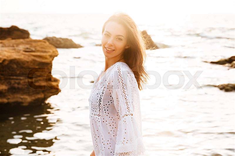 Side view of confident woman in light summer dress walks on beach and looking at the camera, stock photo