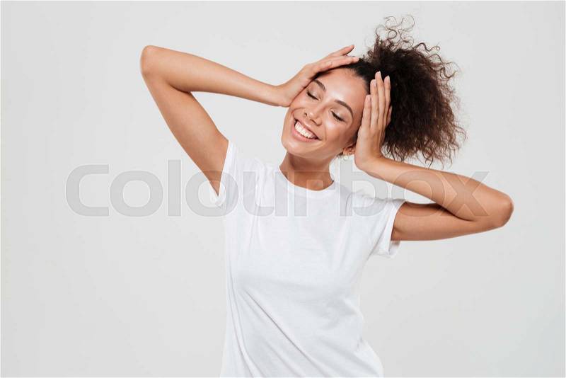 Pleased woman with closed eyes holding hands on head over gray background, stock photo