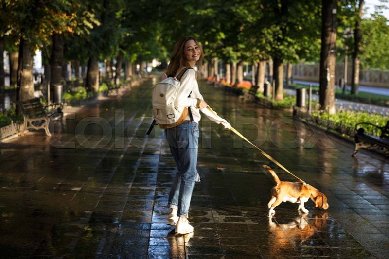 Full length back view image of happy brunette woman in autumn clothes walks with dog in park and looking at the camera, stock photo