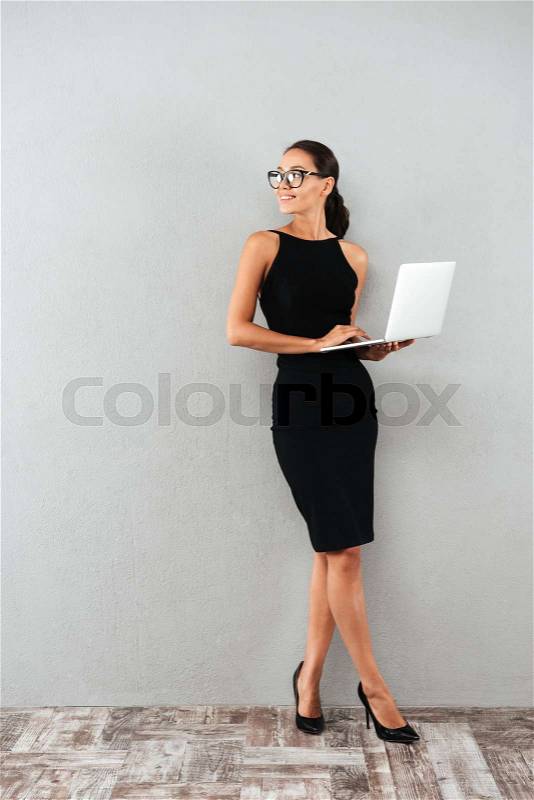 Full length portrait of a beautiful happy businesswoman in black dress and eyeglasses holding laptop computer and looking away at copy space while standing isolated over gray background, stock photo