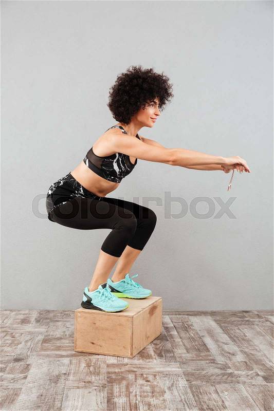 Vertical image of smiling fitness woman jumping on box over gray background, stock photo