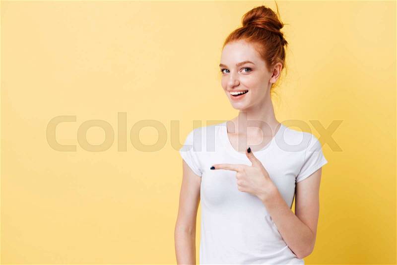 Happy ginger woman in t-shirt pointing away and looking at the camera over yellow background, stock photo