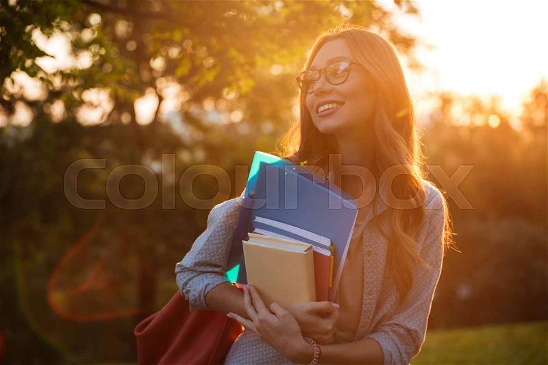 Smiling brunette woman in eyeglasses hugging books and looking away in park, stock photo
