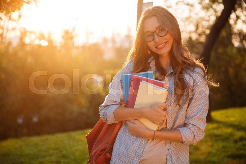 Smiling brunette woman in eyeglasses hugging books and looking at the camera in park, stock photo