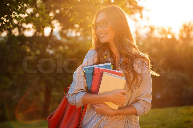 Happy brunette woman in eyeglasses hugging books and looking away in park, stock photo