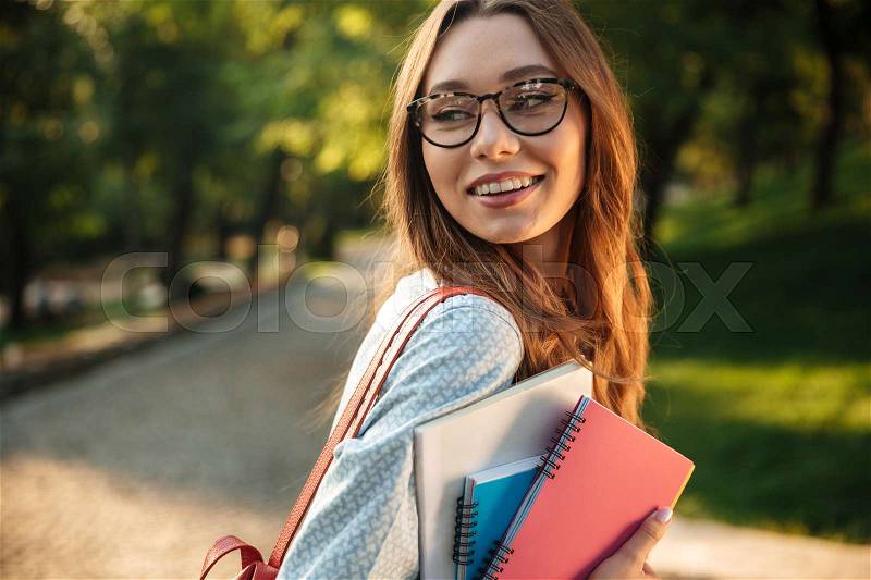 Side view of smiling brunette woman in eyeglasses posing in park with books and looking back, stock photo