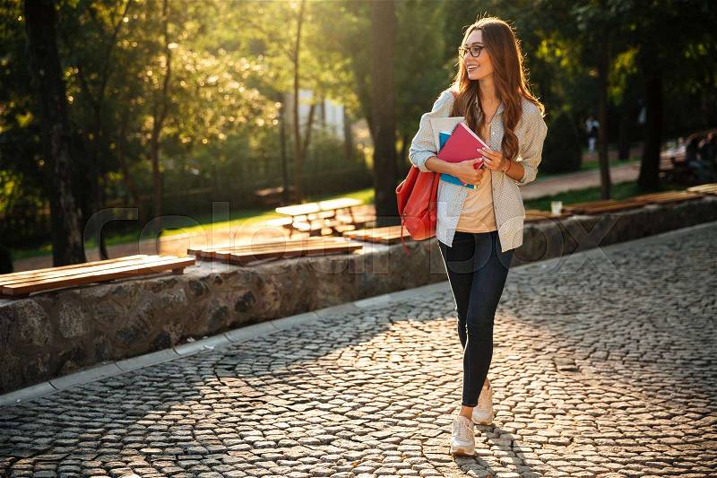 Full length image of happy brunette woman walking with books in park and looking away, stock photo