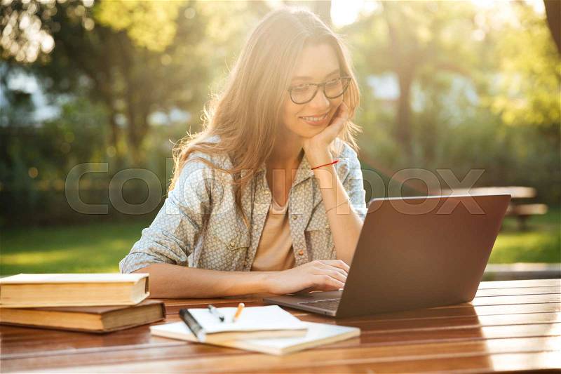 Smiling brunette woman in eyeglasses sitting by the table in park with books and using laptop computer, stock photo
