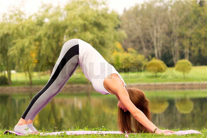Young woman doing yoga asana Downward Facing Dog in the park on the riverside. Adho Mukha Shvanasana, stock photo