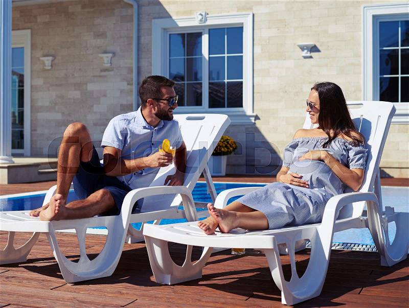 Mid shot of married couple talking while resting on lounge chairs near pool in front of house, stock photo