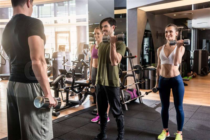 Low-angle view of three young people smiling while alternating dumbbell bicep curl exercise during group workout at the gym, stock photo