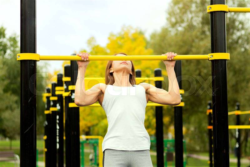 Young athletic fitness woman working out at outdoor gym doing pull ups at sunrise, stock photo