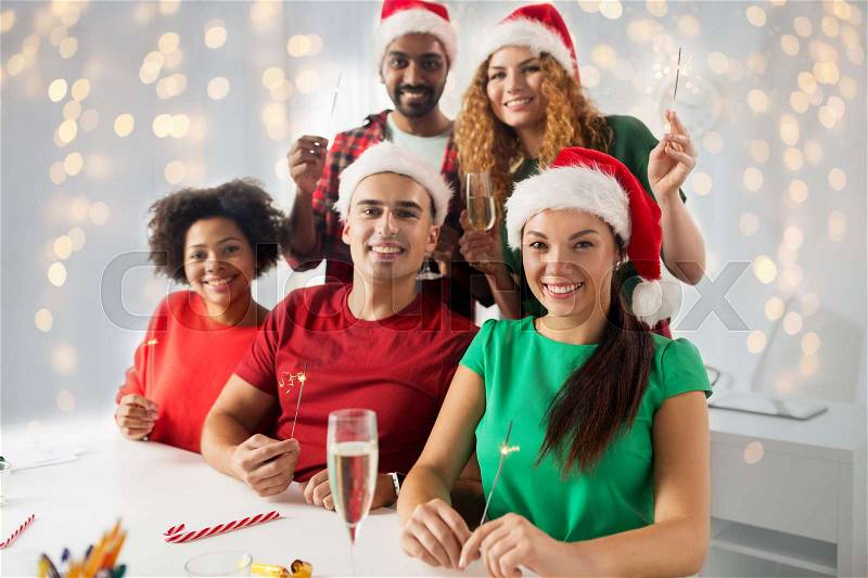 Christmas, celebration and holidays concept - happy team in santa hats with sparklers and non-alcoholic sparkling wine at corporate office party over lights background, stock photo