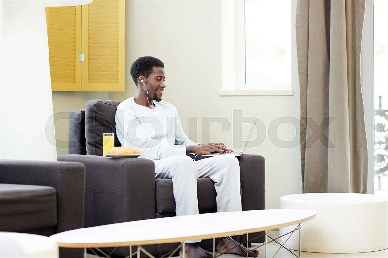 Handsome African American man enjoying lazy weekend at home: he sitting on cozy armchair and playing computer game, interior of modern living room on background, stock photo