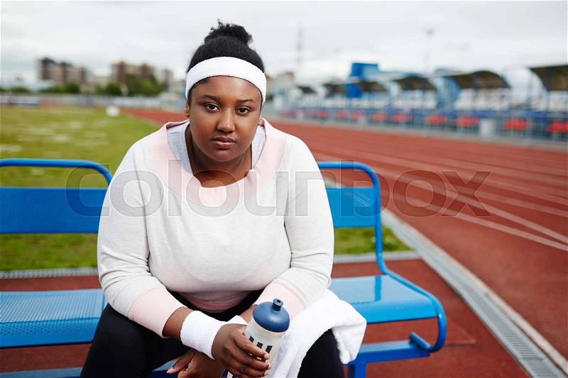 Tired oversized girl in sportswear sitting on bench with towel and bottle of water, stock photo