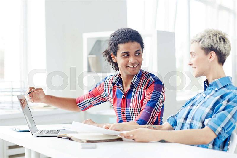 Smiling young interior designers wearing checked shirts working together on joint project while sitting at desk in spacious open plan office, handsome mixed race man pointing at laptop screen, stock photo
