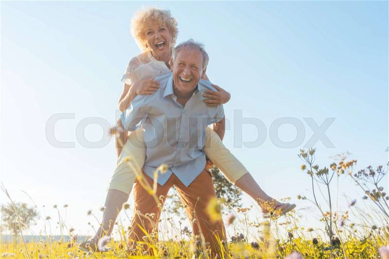 Low-angle view portrait of a happy senior man laughing while carrying his partner on his back, in a sunny day of summer in the countryside, stock photo