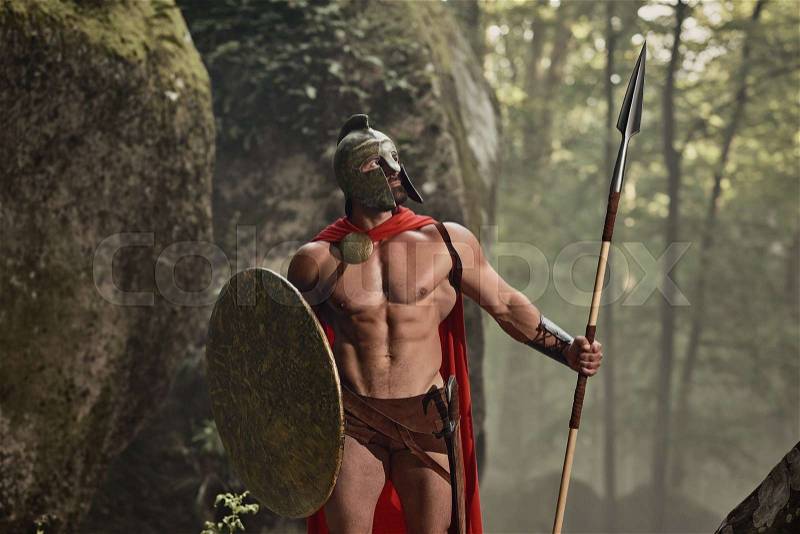 Sportive man with great muscles wearing armor of ancient gladiator and posing with arms on background of rocks and woods, stock photo