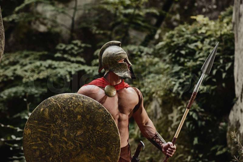 Muscular man wearing outfit of ancient gladiator holding long spear and shield and posing at camera looking away. Spartan, stock photo