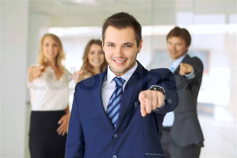 Smiling businessman in office with colleagues in the background. Pointing by finger into the camera, stock photo