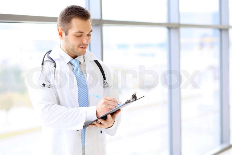 Smiling doctor filling up medical history form while standing near big window, stock photo
