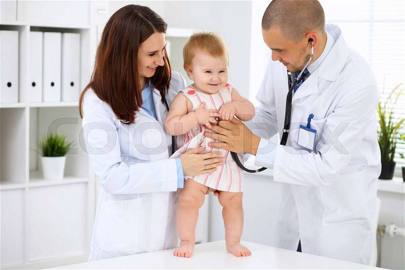 Happy cute baby at health exam at doctor's office. Toddler girl is standing while have been keeping by two doctors, stock photo