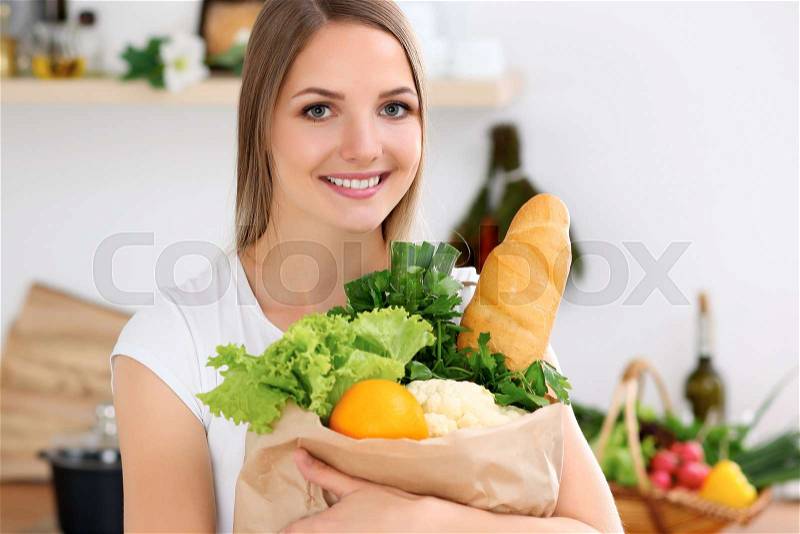 Young cheerful smiling woman is ready for cooking in a kitchen. Housewife is holding big paper bag full of fresh vegetables and fruits and looking at the camera, stock photo