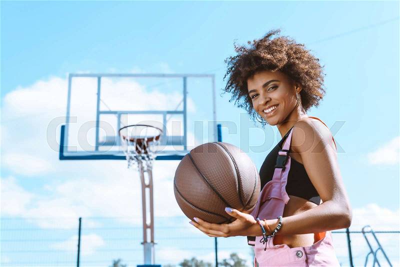 African-american woman in sports bra and pink overalls holding a basketball ball at sports court, stock photo