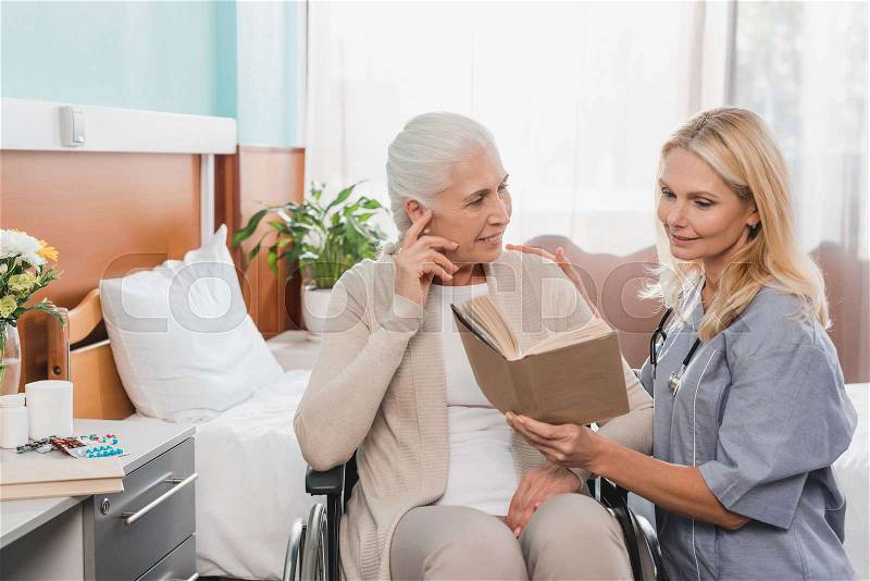 Smiling nurse reading book to senior patient with hearing aid sitting in wheelchair , stock photo