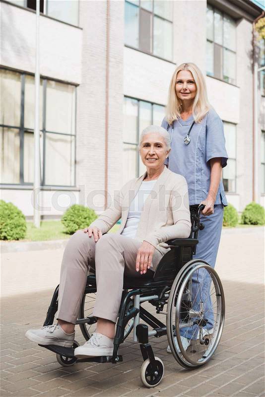 Happy senior woman in wheelchair and nurse smiling at camera, stock photo