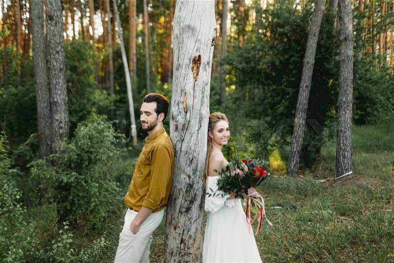 Bride and groom lean on the tree from different sides. Newlyweds are walking in the forest. Artwork,, stock photo
