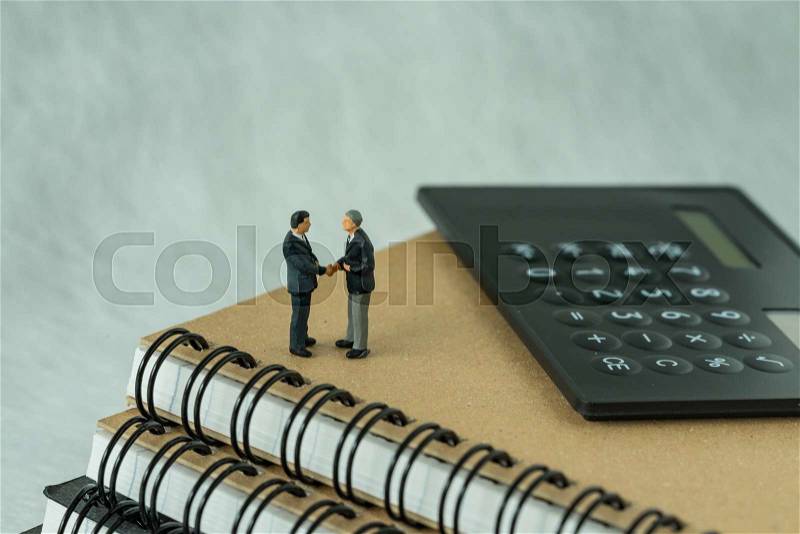 Miniature people: Small figure businessmen handshaking and standing on notebook with calculator as business agreement concept, stock photo