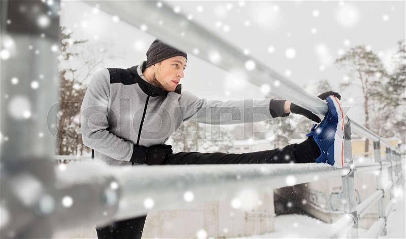 Fitness, sport, people, exercising and healthy lifestyle concept - young man stretching leg and warming up at fence in winter, stock photo