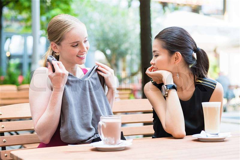 Asian fashionable young woman showing to her best friend a new trendy purchase while sitting down at a trendy cafeteria downtown, stock photo