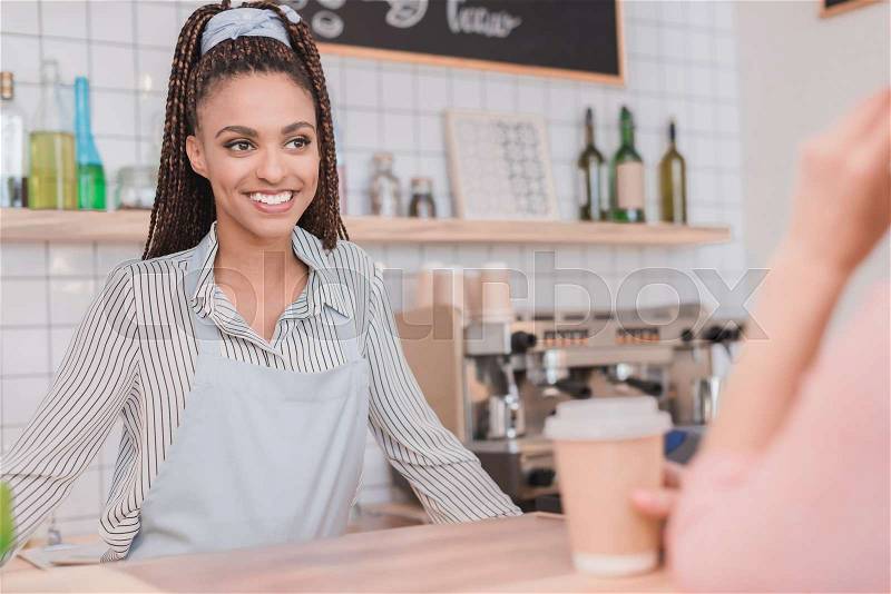 Smiling african american barista standing behind the counter at coffee shop and looking at client, stock photo