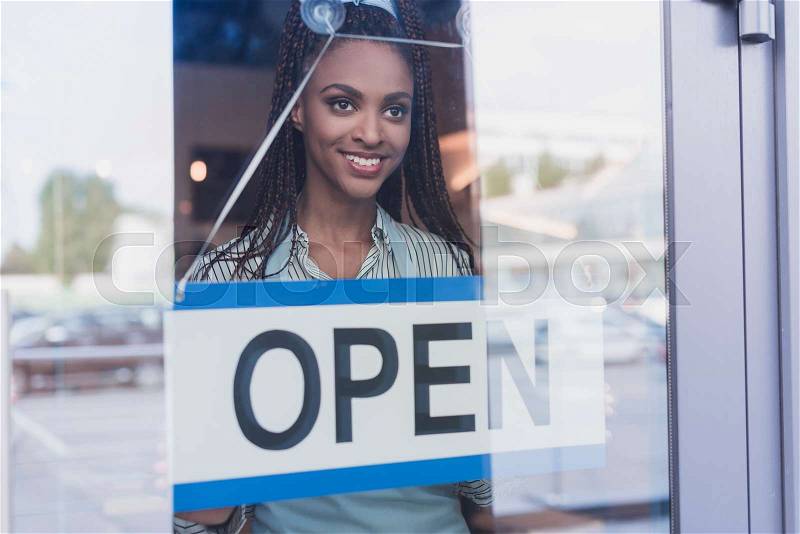 Young african american woman hanging an open sign on a glass door, stock photo