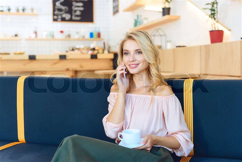 Beautiful young woman holding cup of coffee and talking on smartphone in cafe , stock photo