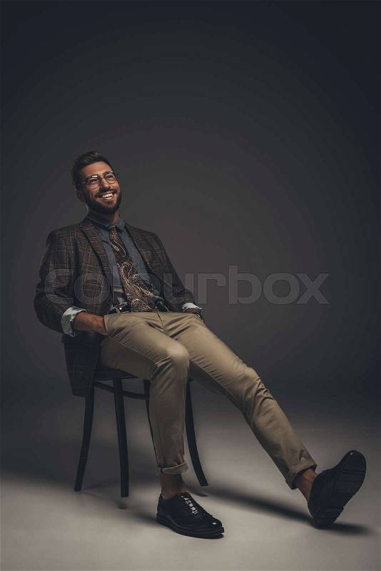 Laughing young man in suit and glasses sitting on chair with hands in pockets, stock photo