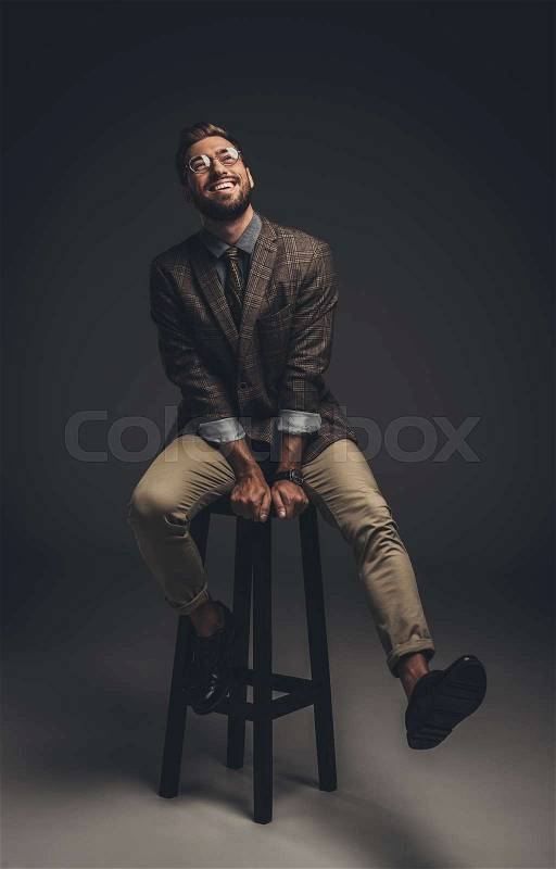 Laughing young man in suit and glasses sitting on bar stool, stock photo