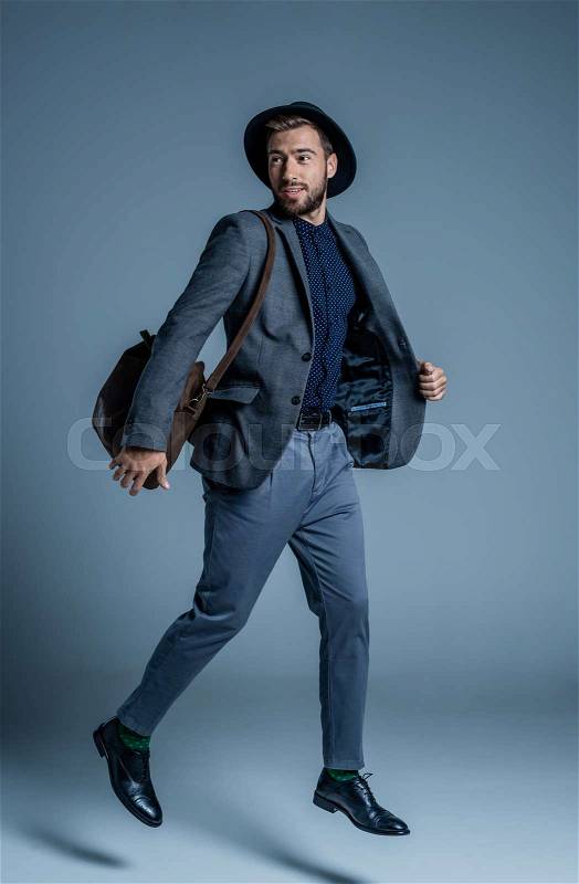 Smiling young man in suit and hat jumping up with leather bag on his shoulder, stock photo