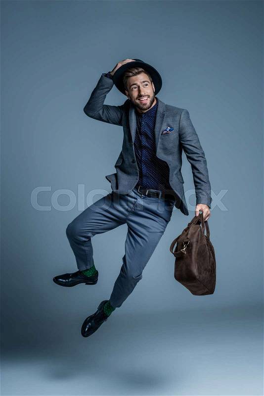 Smiling young man in suit and hat jumping up with leather bag in his hand, stock photo