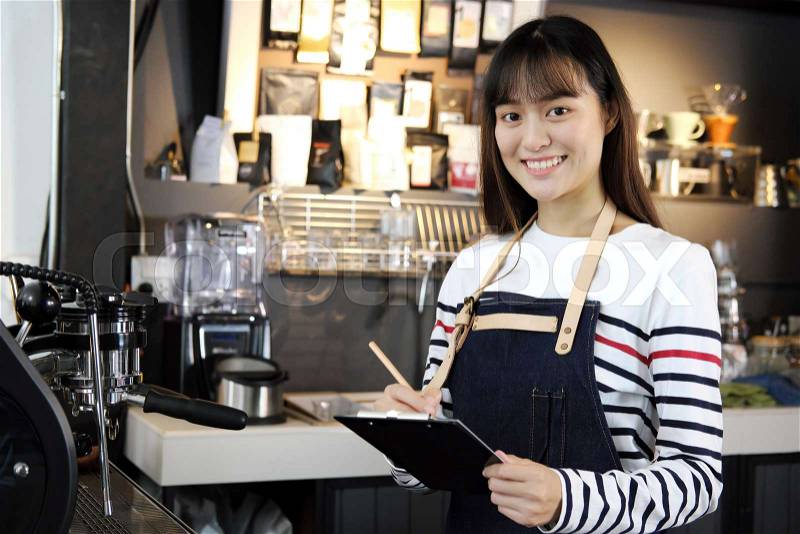Portrait of smiling asian barista listing food. Young asian woman barista in her shop. Cafe restaurant service, food and drink industry concept, stock photo