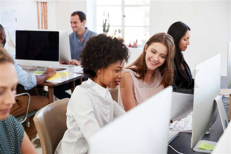 Two happy women discuss work at computer in open plan office, stock photo