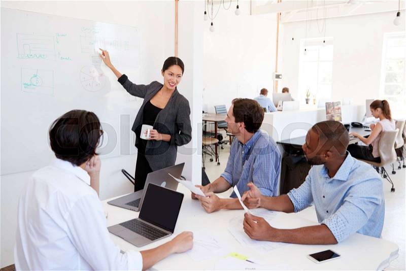 Woman pointing at whiteboard at a meeting in a busy office, stock photo